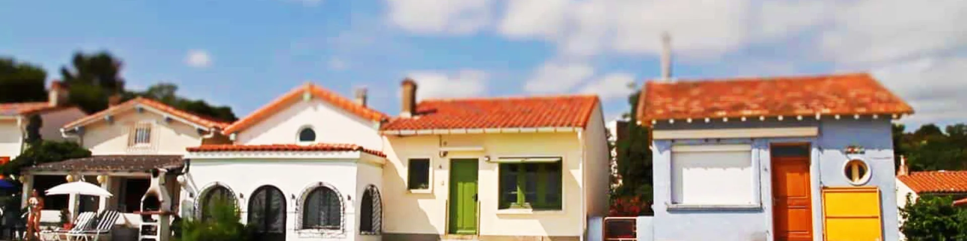 Alignement de petites maisons colorées du Racou à Argelès-sur-Mer, photographiées de face depuis la plage, avec leurs toits en tuiles rouges, façades pastel et volets colorés sous un ciel bleu légèrement nuageux.