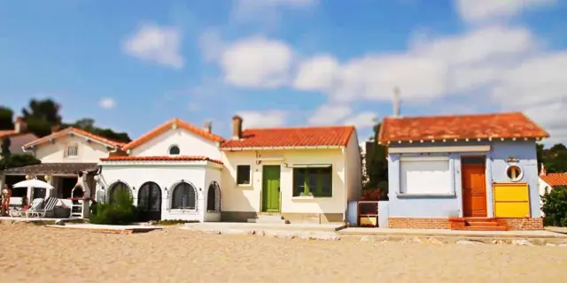 Alignement de petites maisons colorées du Racou à Argelès-sur-Mer, photographiées de face depuis la plage, avec leurs toits en tuiles rouges, façades pastel et volets colorés sous un ciel bleu légèrement nuageux.