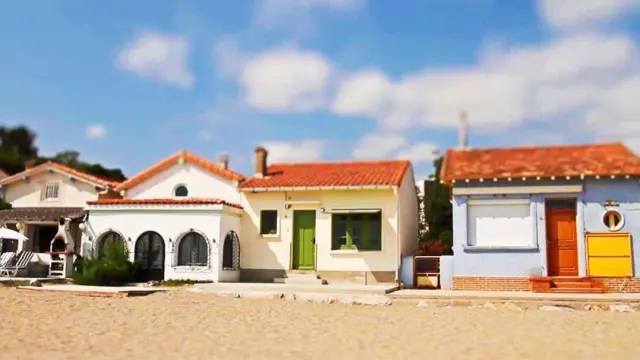 Alignement de petites maisons colorées du Racou à Argelès-sur-Mer, photographiées de face depuis la plage, avec leurs toits en tuiles rouges, façades pastel et volets colorés sous un ciel bleu légèrement nuageux.