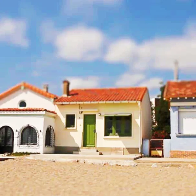 Alignement de petites maisons colorées du Racou à Argelès-sur-Mer, photographiées de face depuis la plage, avec leurs toits en tuiles rouges, façades pastel et volets colorés sous un ciel bleu légèrement nuageux.