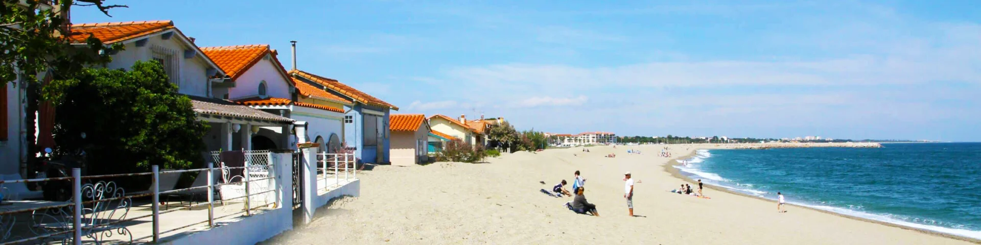 Vue depuis la plage du Racou à Argelès-sur-Mer, montrant les petites maisons aux toits en tuiles rouges alignées le long du sable blond, avec quelques vacanciers profitant du bord de mer sous un ciel bleu clair.