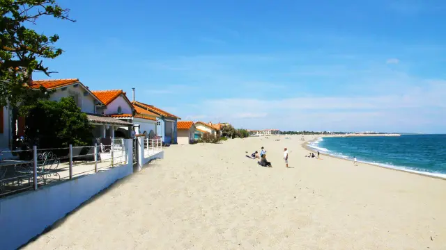 Vue depuis la plage du Racou à Argelès-sur-Mer, montrant les petites maisons aux toits en tuiles rouges alignées le long du sable blond, avec quelques vacanciers profitant du bord de mer sous un ciel bleu clair.