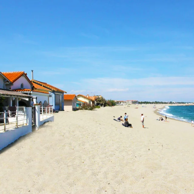 Vue depuis la plage du Racou à Argelès-sur-Mer, montrant les petites maisons aux toits en tuiles rouges alignées le long du sable blond, avec quelques vacanciers profitant du bord de mer sous un ciel bleu clair.