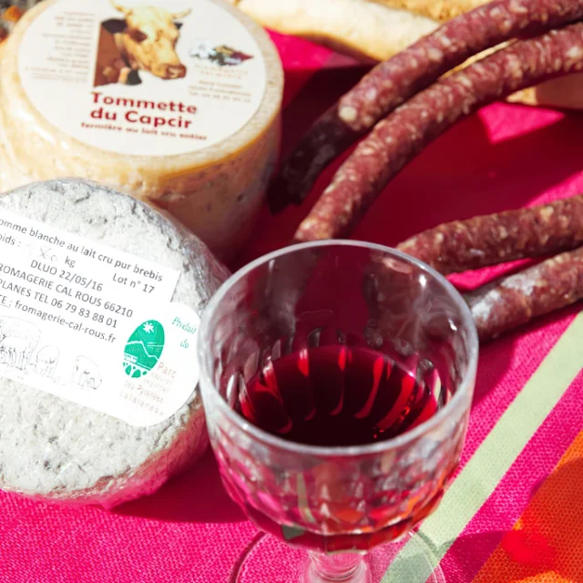 Détail d’un petit déjeuner montagnard avec fromages fermiers, saucisson, pain et verre de vin rouge, posés sur une nappe catalane colorée.