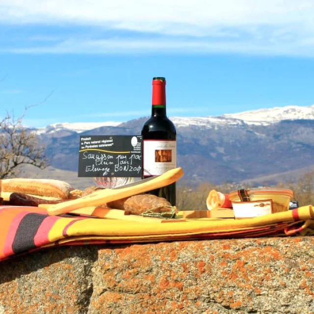 Petit-déjeuner montagnard sur une nappe catalane avec pain, fromage, charcuterie et vin rouge, face aux montagnes enneigées du site des Bouillouses.