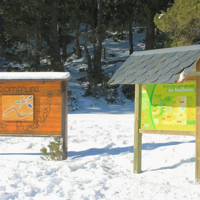 Deux panneaux en bois installés dans la neige à l’entrée du site classé des Bouillouses : l’un indiquant « Commune Bolquère Pyrénées 2000 » et l’autre présentant la carte d’accueil verte avec la mention « Bienvenue sur le site classé des Bouillouses », sur fond de forêt enneigée.