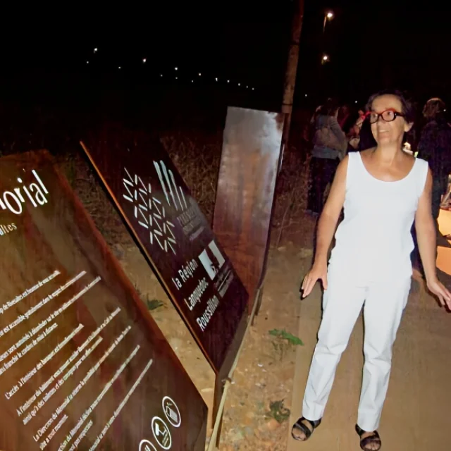 Femme souriante en tenue blanche se tenant devant les panneaux d’entrée du Mémorial du Camp de Rivesaltes, de nuit, entourée d’autres visiteurs.