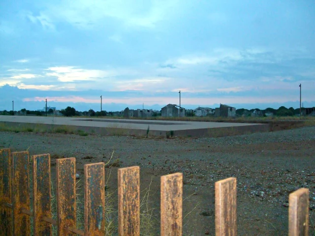 Vue du Mémorial du Camp de Rivesaltes au crépuscule, avec une clôture rouillée au premier plan et les anciens bâtiments du camp à l’horizon sous un ciel bleu rosé.