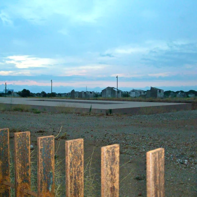 Vue du Mémorial du Camp de Rivesaltes au crépuscule, avec une clôture rouillée au premier plan et les anciens bâtiments du camp à l’horizon sous un ciel bleu rosé.