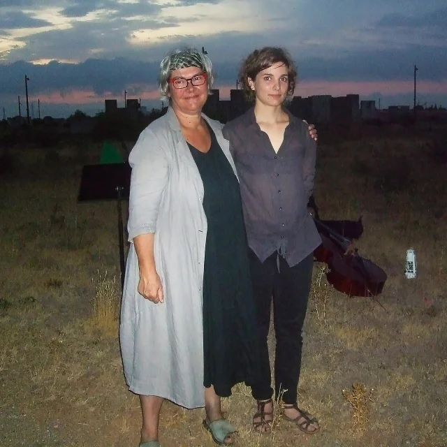 Deux femmes debout côte à côte au Mémorial du Camp de Rivesaltes, photographiées à la tombée du jour sur un terrain herbeux, avec un ciel aux teintes bleutées et rosées en arrière-plan.