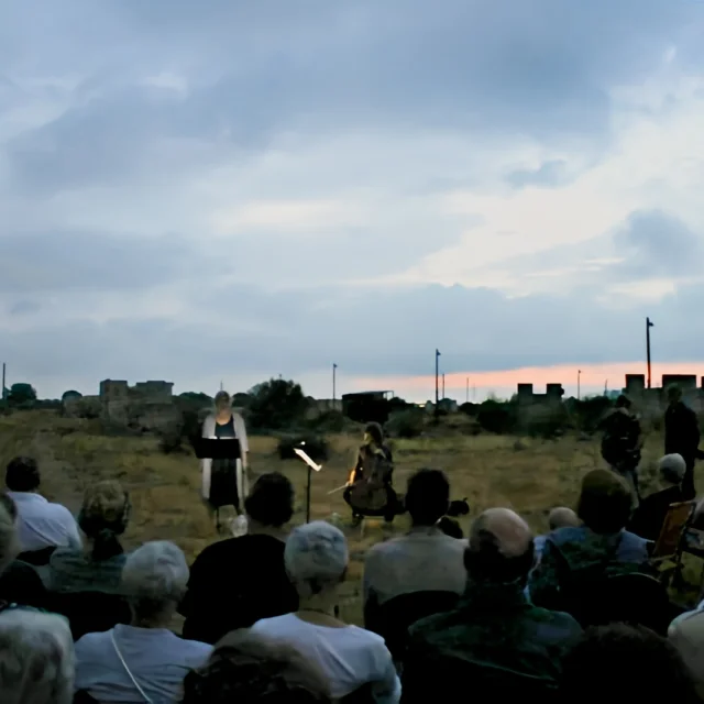 Lecture publique accompagnée de musique au Mémorial du Camp de Rivesaltes, en plein air au crépuscule, devant un public assis face à la scène.