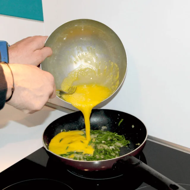 Préparation d’une omelette aux asperges dans une poêle, un mélange d’œufs battus est versé sur des asperges cuites.