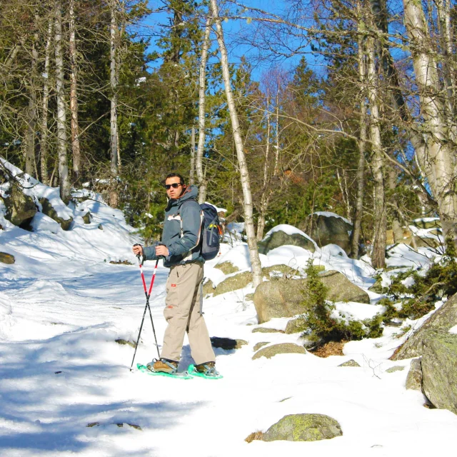 Homme équipé de raquettes, d’un sac à dos et de bâtons de marche, avançant sur un sentier enneigé à travers la forêt des Bouillouses, entouré de pins et de rochers sous un ciel bleu.
