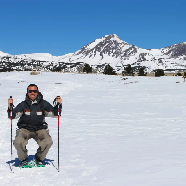 Homme assis dans la neige, souriant, équipé de raquettes et de bâtons de randonnée, face aux sommets enneigés des Bouillouses sous un ciel bleu éclatant.