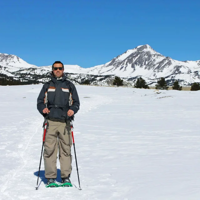 Homme debout en tenue de randonnée hivernale, équipé de raquettes et de bâtons, sur une vaste plaine enneigée du site des Bouillouses. À l’arrière-plan, les montagnes du Capcir se dressent sous un ciel bleu éclatant.