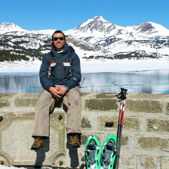 Homme assis sur le muret du barrage des Bouillouses, vêtu d’une tenue de montagne, avec des raquettes et des bâtons posés à côté de lui. En arrière-plan, le lac gelé et les sommets enneigés du Capcir sous un ciel bleu.