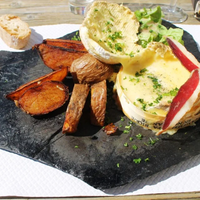 Plat de camembert rôti au four servi sur une ardoise, accompagné de pommes de terre dorées, salade verte et tranches de jambon sec, en terrasse du refuge des Bouillouses.