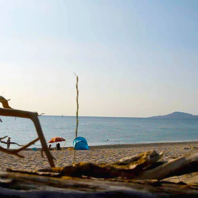 Vue sur la plage de la Réserve Naturelle du Mas Larrieu au coucher du soleil, avec des vacanciers installés sous une tente et un parapluie coloré, la mer calme et les collines à l’horizon.