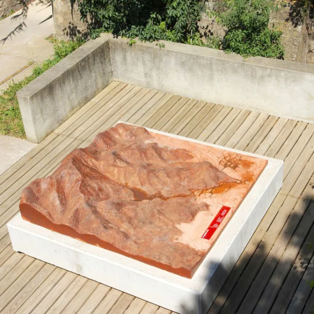 Vue aérienne d’une maquette topographique en relief représentant le site de Paulilles. Installée sur une terrasse en bois, la maquette de couleur ocre est entourée de murets en béton et de végétation.