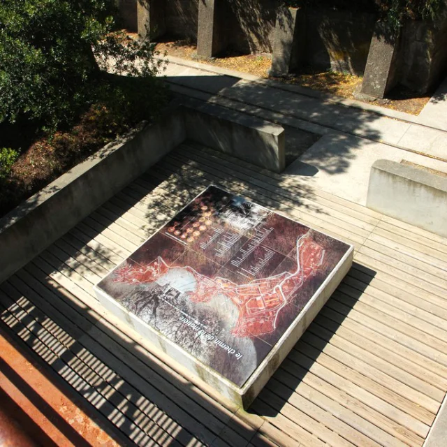 Vue plongeante sur une table d’orientation représentant le plan du site des anciennes dynamiteries Alfred Nobel à Paulilles. Le panneau, installé sur une terrasse en bois encadrée de murets de béton, est partiellement ombragé par la végétation environnante.