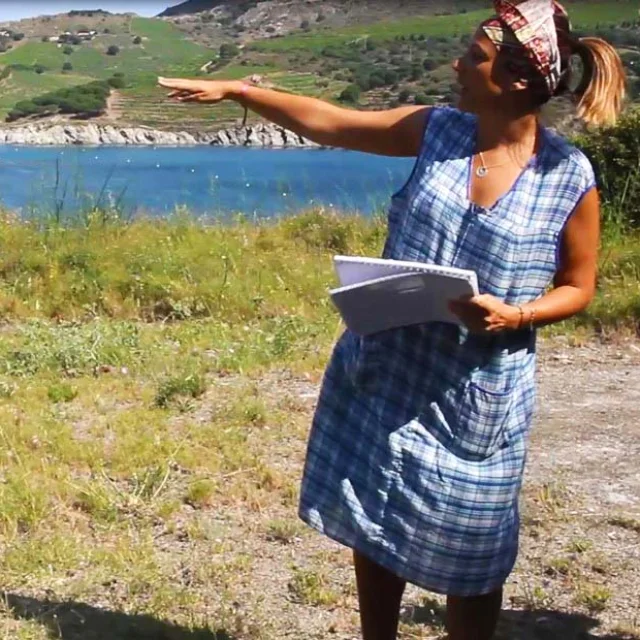Une femme en robe à carreaux bleus, carnet en main, désigne du bras la baie de Paulilles en arrière-plan, avec la mer turquoise et les collines verdoyantes couvertes de vignes sous un ciel clair.