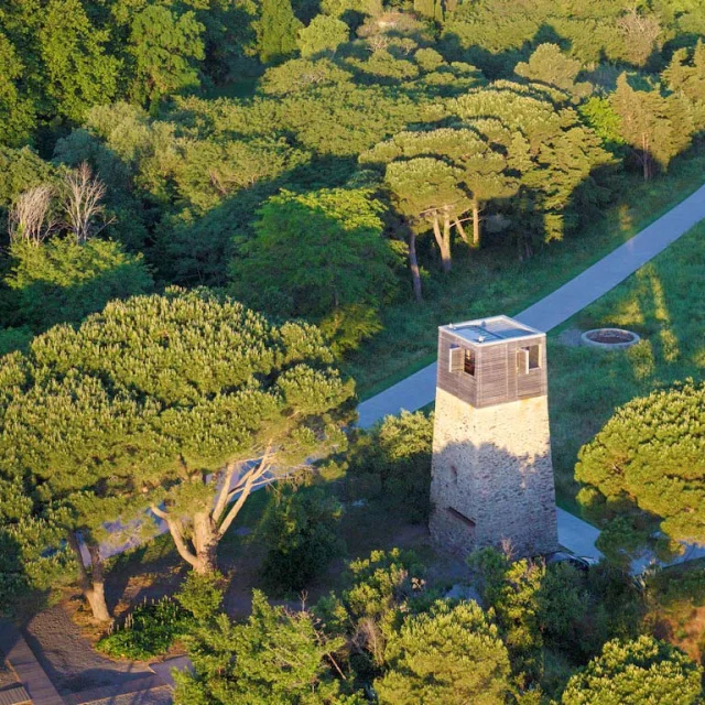 Vue aérienne de la tour de guet du site de Paulilles, entourée d’une dense végétation méditerranéenne. La tour en pierre, surmontée d’un observatoire en bois, se dresse au bord d’un chemin qui serpente à travers les pins et les collines verdoyantes.