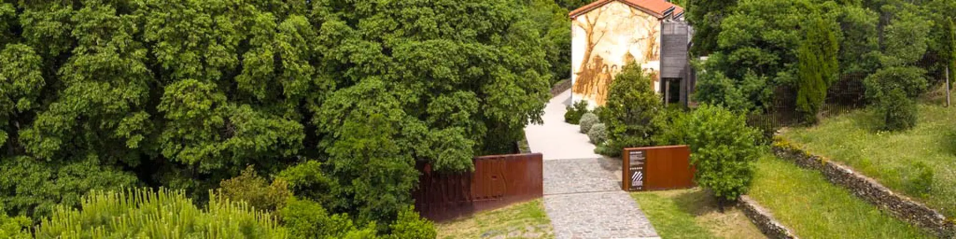 Vue aérienne de l’entrée du site des anciennes dynamiteries Alfred Nobel à Paulilles. Un chemin pavé traverse un espace verdoyant menant à un bâtiment restauré, orné d’une fresque murale représentant des ouvriers du site, entouré d’arbres et de végétation dense.