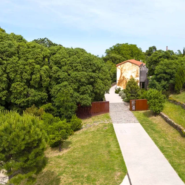 Vue aérienne de l’entrée du site des anciennes dynamiteries Alfred Nobel à Paulilles. Un chemin pavé traverse un espace verdoyant menant à un bâtiment restauré, orné d’une fresque murale représentant des ouvriers du site, entouré d’arbres et de végétation dense.