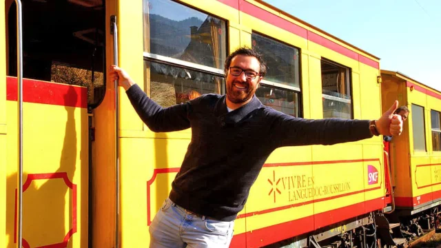Un homme souriant s’accroche à la porte ouverte du Train Jaune, les bras écartés, pouce levé, exprimant la joie de voyager à bord du célèbre train jaune et rouge des Pyrénées-Orientales.