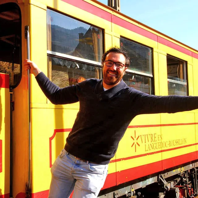 Un homme souriant s’accroche à la porte ouverte du Train Jaune, les bras écartés, pouce levé, exprimant la joie de voyager à bord du célèbre train jaune et rouge des Pyrénées-Orientales.