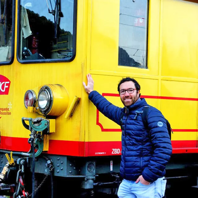 Un homme souriant pose devant la motrice jaune et rouge du Train Jaune des Pyrénées-Orientales, stationné à quai. On distingue les logos SNCF et Région Languedoc-Roussillon sur la locomotive.