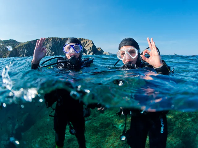 Deux plongeurs équipés de combinaisons, masques et bouteilles d’air sont à la surface de la mer, faisant le signe “OK” avec la main. En arrière-plan, on distingue les falaises rocheuses de la Côte Vermeille sous un ciel bleu clair.