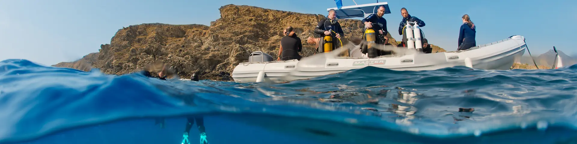 Vue mi-aérienne mi-sous-marine d’un groupe de plongeurs à bord d’un bateau pneumatique blanc, équipé de bouteilles de plongée. Sous la surface, un plongeur est visible avec ses palmes turquoise, prêt à explorer les fonds marins près d’une côte rocheuse.