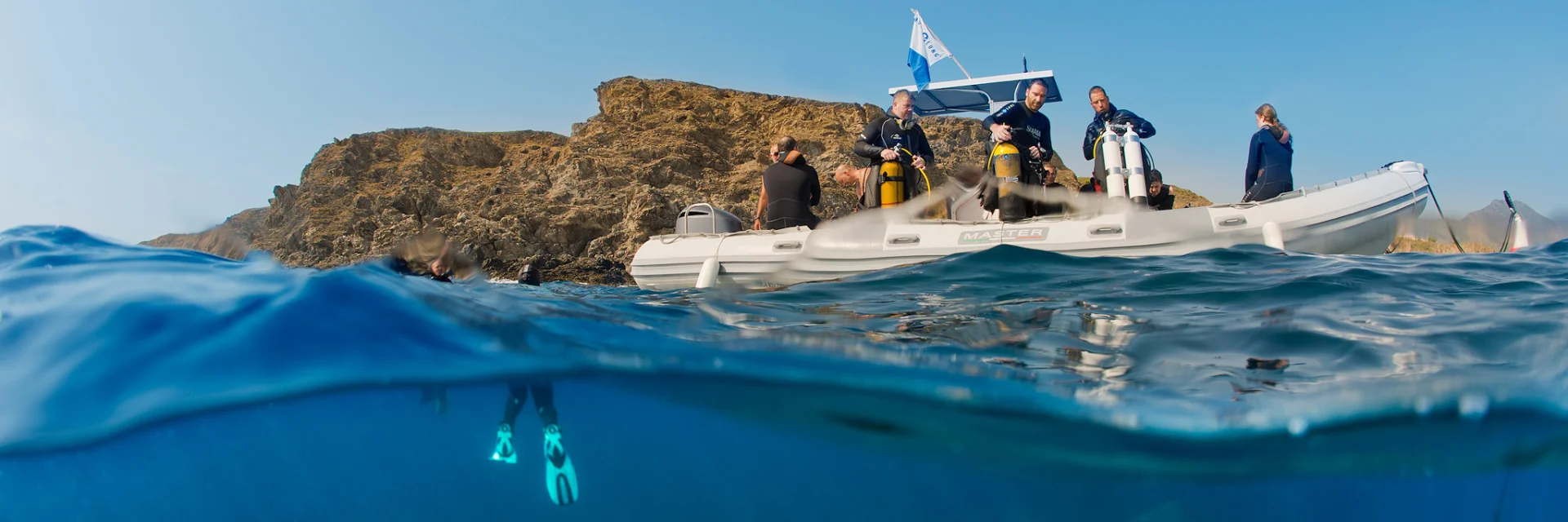 Vue mi-aérienne mi-sous-marine d’un groupe de plongeurs à bord d’un bateau pneumatique blanc, équipé de bouteilles de plongée. Sous la surface, un plongeur est visible avec ses palmes turquoise, prêt à explorer les fonds marins près d’une côte rocheuse.
