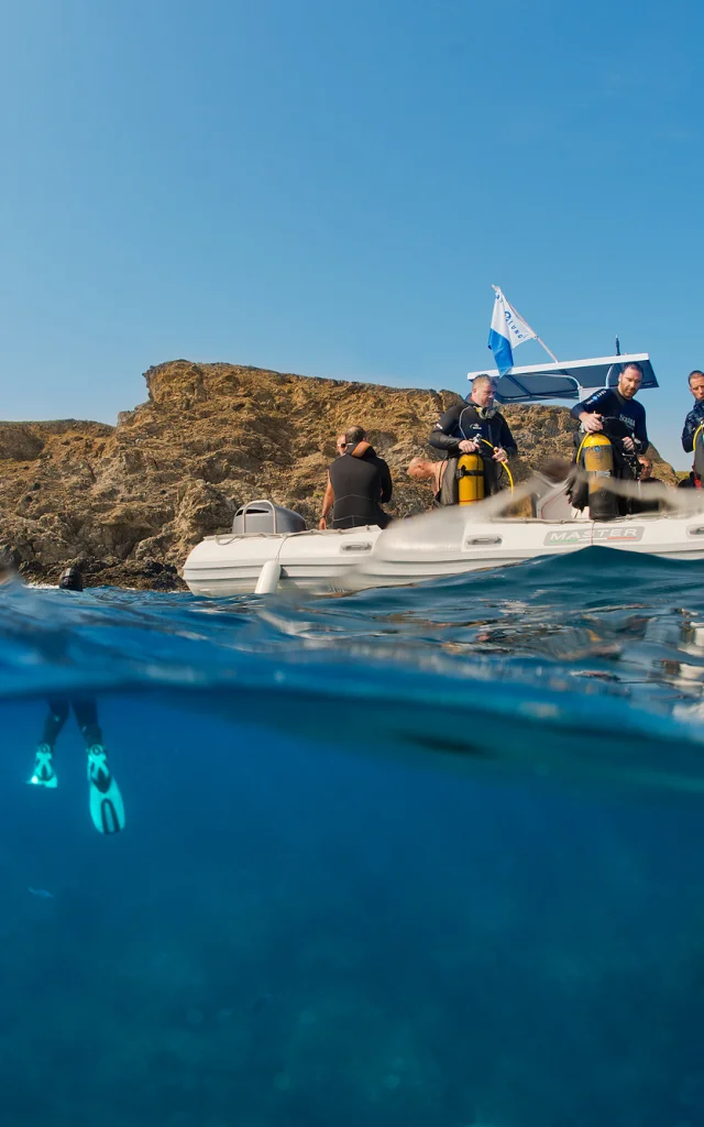 Vue mi-aérienne mi-sous-marine d’un groupe de plongeurs à bord d’un bateau pneumatique blanc, équipé de bouteilles de plongée. Sous la surface, un plongeur est visible avec ses palmes turquoise, prêt à explorer les fonds marins près d’une côte rocheuse.