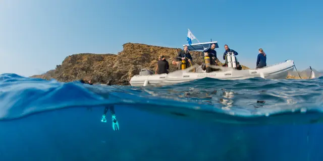 Vue mi-aérienne mi-sous-marine d’un groupe de plongeurs à bord d’un bateau pneumatique blanc, équipé de bouteilles de plongée. Sous la surface, un plongeur est visible avec ses palmes turquoise, prêt à explorer les fonds marins près d’une côte rocheuse.