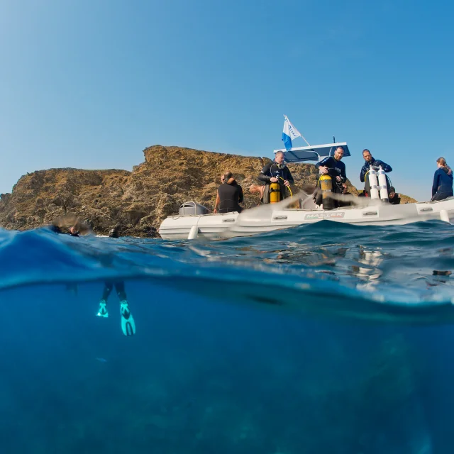 Vue mi-aérienne mi-sous-marine d’un groupe de plongeurs à bord d’un bateau pneumatique blanc, équipé de bouteilles de plongée. Sous la surface, un plongeur est visible avec ses palmes turquoise, prêt à explorer les fonds marins près d’une côte rocheuse.