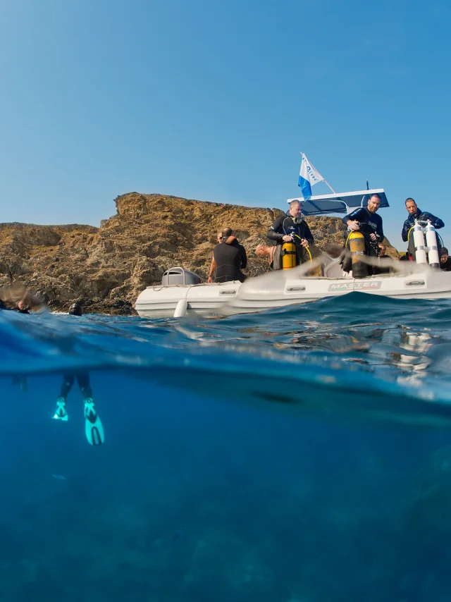 Vue mi-aérienne mi-sous-marine d’un groupe de plongeurs à bord d’un bateau pneumatique blanc, équipé de bouteilles de plongée. Sous la surface, un plongeur est visible avec ses palmes turquoise, prêt à explorer les fonds marins près d’une côte rocheuse.