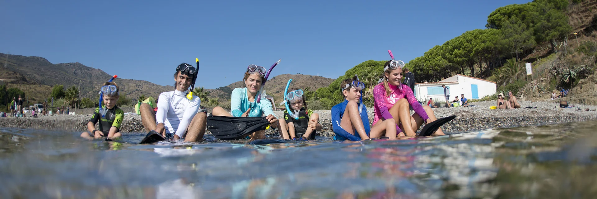 Un groupe composé de deux adultes et quatre enfants, équipés de masques, tubas et palmes, est assis au bord de l’eau sur une plage de galets. Ils sourient et ajustent leur matériel avant d’entrer dans la mer, sous un ciel bleu et face à la côte méditerranéenne.
