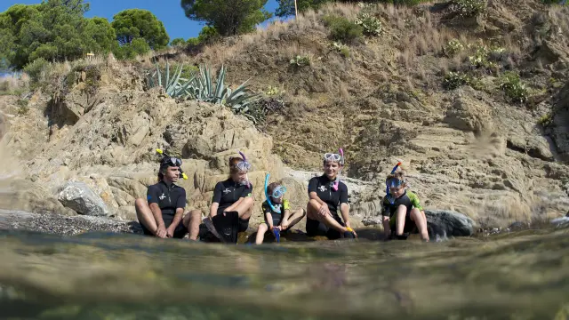 Un groupe de cinq personnes, adultes et enfants, assis sur les rochers au bord de l’eau, portent des combinaisons de plongée, masques et tubas. Ils se préparent pour une activité de snorkeling le long d’une côte rocheuse bordée de végétation méditerranéenne.