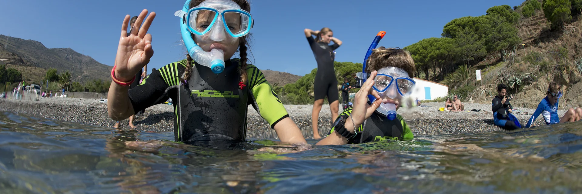 Deux enfants équipés de combinaisons de plongée, de masques et de tubas sont dans l’eau près du rivage. Ils font un signe de la main “ok” en direction de l’objectif. En arrière-plan, on aperçoit d’autres personnes sur la plage et la végétation méditerranéenne.