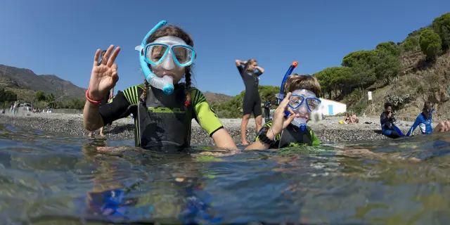 Deux enfants équipés de combinaisons de plongée, de masques et de tubas sont dans l’eau près du rivage. Ils font un signe de la main “ok” en direction de l’objectif. En arrière-plan, on aperçoit d’autres personnes sur la plage et la végétation méditerranéenne.