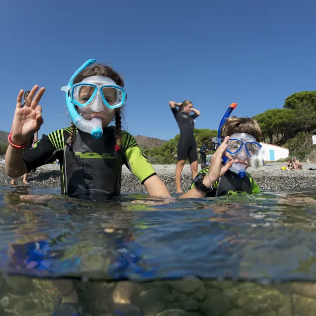 Deux enfants équipés de combinaisons de plongée, de masques et de tubas sont dans l’eau près du rivage. Ils font un signe de la main “ok” en direction de l’objectif. En arrière-plan, on aperçoit d’autres personnes sur la plage et la végétation méditerranéenne.