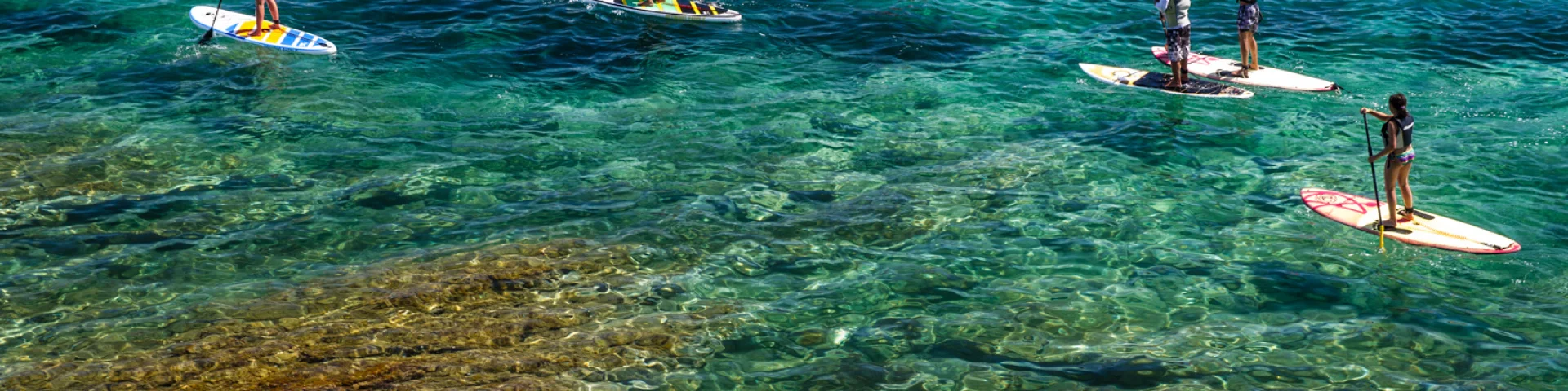 Groupe de personnes pratiquant le paddle sur une mer turquoise et transparente, au-dessus d’un fond rocheux visible.