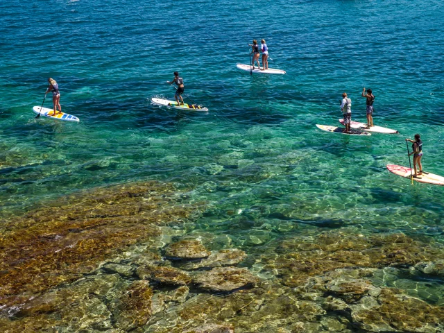Groupe de personnes pratiquant le paddle sur une mer turquoise et transparente, au-dessus d’un fond rocheux visible.