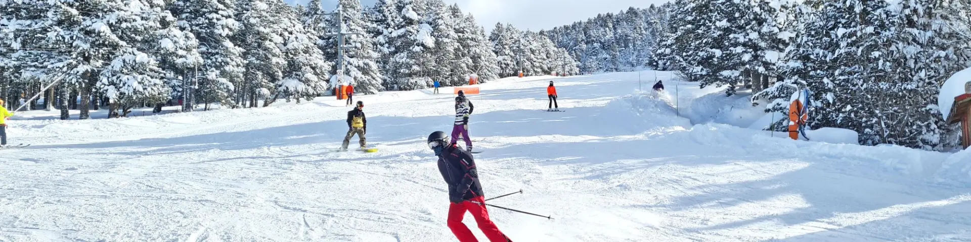 Skieurs et snowboarders sur une piste enneigée bordée de sapins chargés de neige sous un ciel bleu partiellement nuageux.