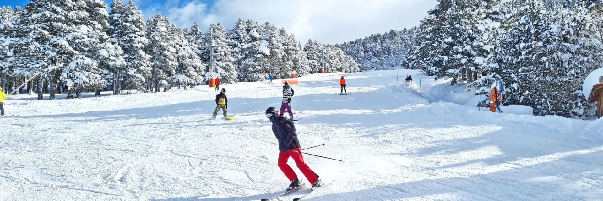 Skieurs et snowboarders sur une piste enneigée bordée de sapins chargés de neige sous un ciel bleu partiellement nuageux.
