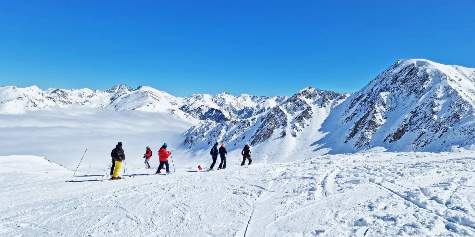 Groupe de skieurs sur une piste enneigée dominant une mer de nuages, entourée de hauts sommets sous un ciel bleu.