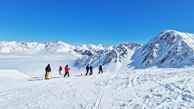 Groupe de skieurs sur une piste enneigée dominant une mer de nuages, entourée de hauts sommets sous un ciel bleu.