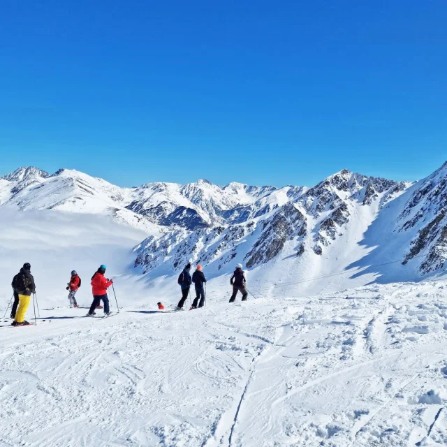 Groupe de skieurs sur une piste enneigée dominant une mer de nuages, entourée de hauts sommets sous un ciel bleu.